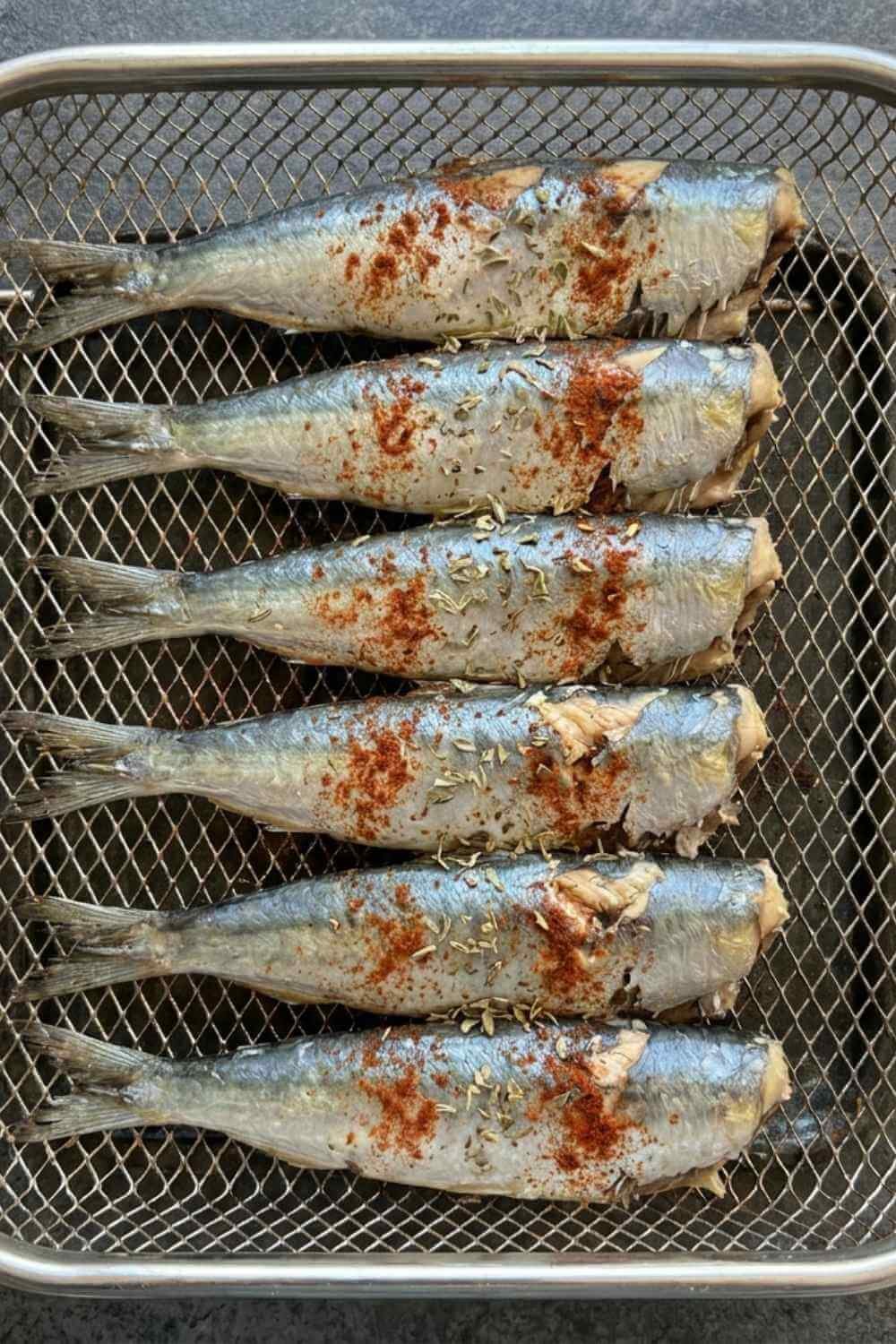 Canned sardines arranged in a single layer in an air fryer basket, dusted with smoked paprika and oregano
